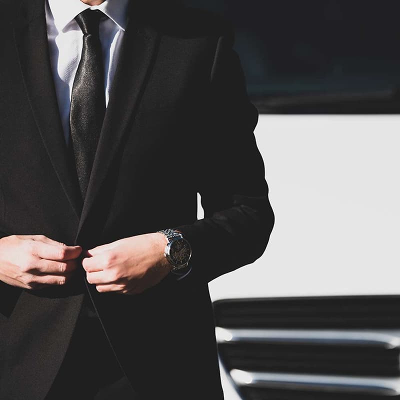 Costume noir & Élégance : L'homme d'affaires prêt. Homme élégant en costume noir, cravate noire et chemise blanche, boutonnant sa veste. Montre-bracelet visible. Arrière-plan de limousine.